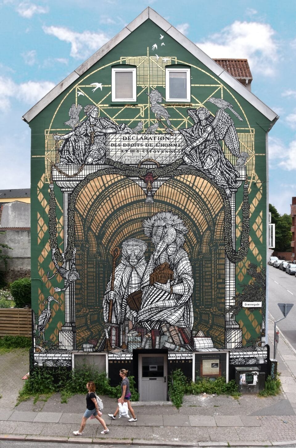 a mural with a black and white stenciled depiction of a a monkey holding a staff and a bird holding an architectural model. the background is green with gold details. angels are at the top with smaller animals to the sides
