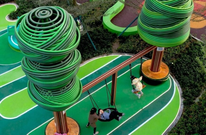Home En 59 an aerial view of part of an elaborately designed playground with blue and green designs on the ground and wire-like sculptural trees holding up bridges and play equipment