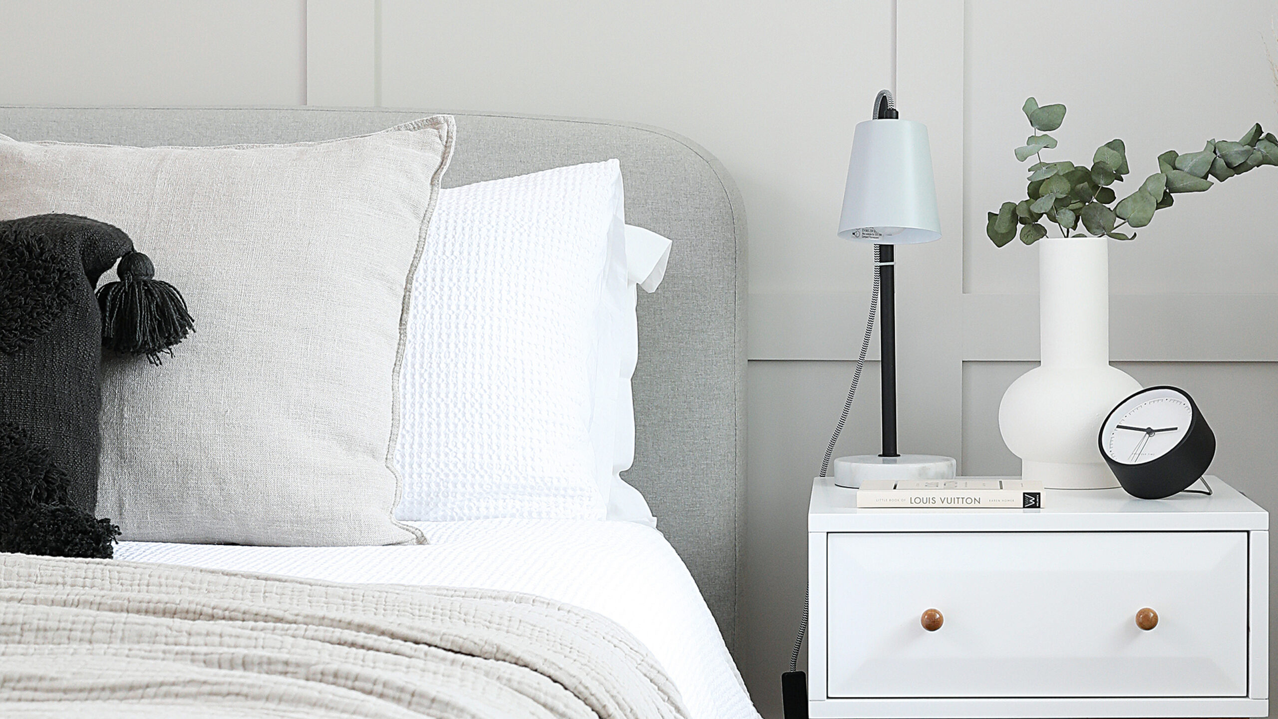 A bed with a grey headboard and white bedding and a white bedside table with a lamp and black alarm clock