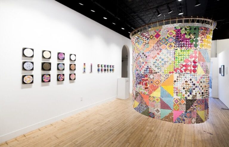 an installation view of a gallery with small artworks on the wall to the left and a suspended textile piece on the right, made from tiny pieces of plastic and paper repurposed into a colorful textile