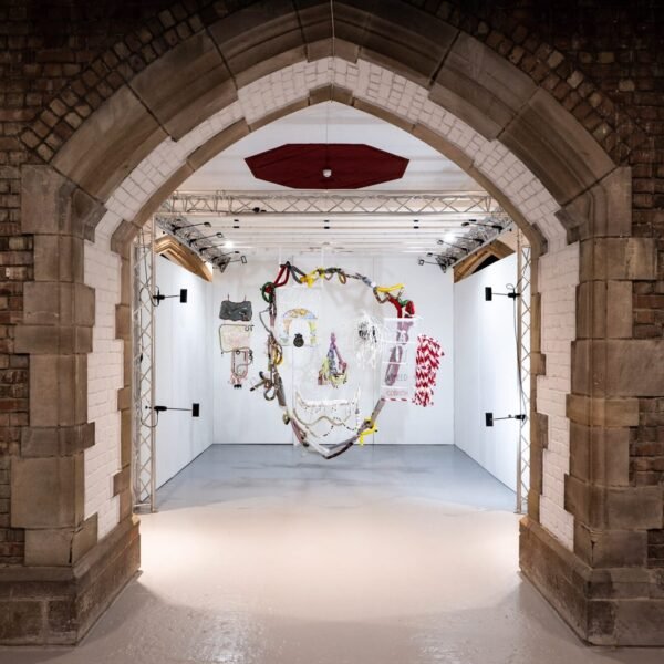 an installation view of an artwork made of various pieces of fabric and found objects, suspended from the ceiling to create a face when viewed straight-on
