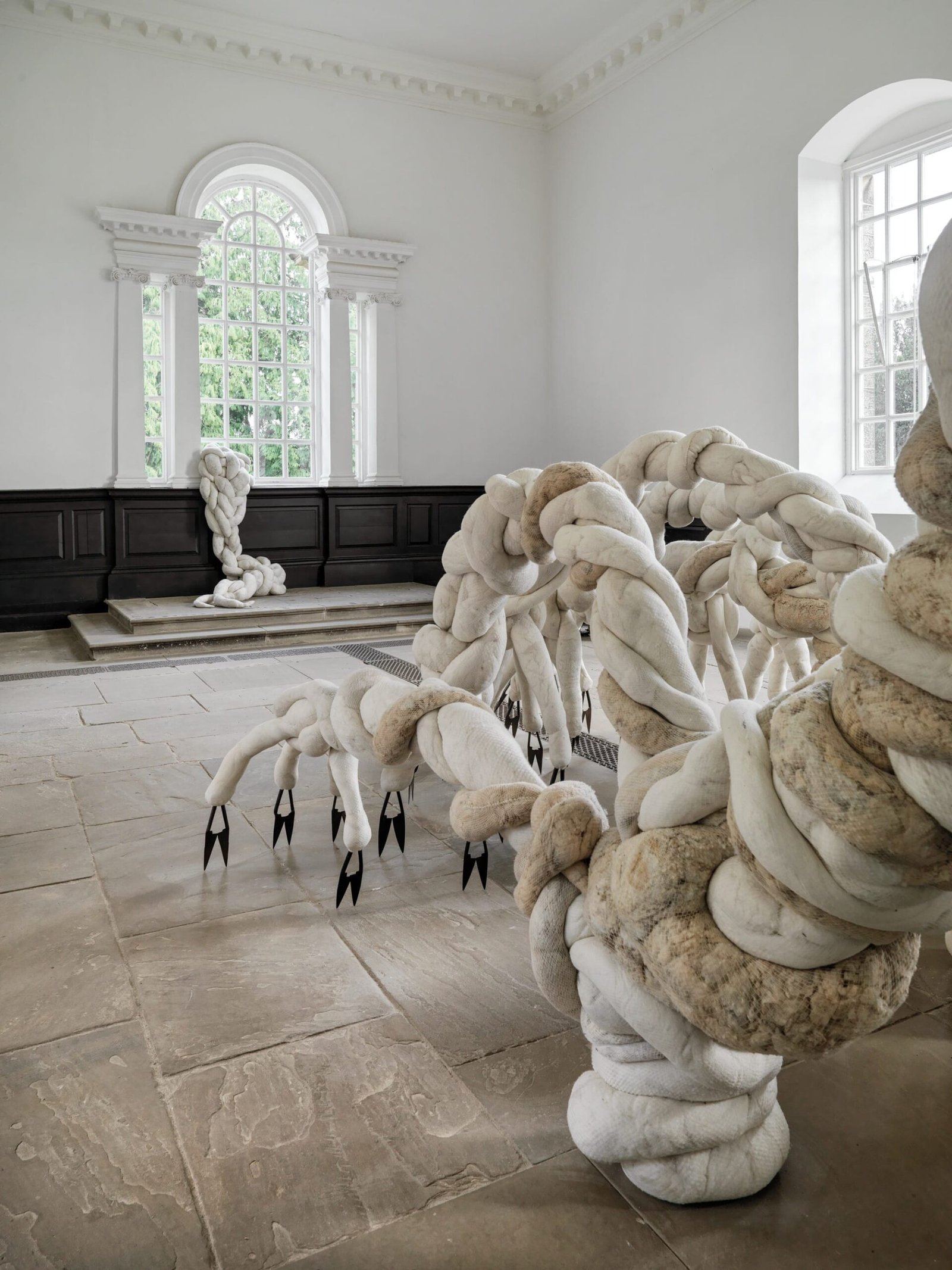 a large-scale installation by Nicola Turner inside of Yorkshire Sculpture Park's 18th-century chapel gallery space of a textile form that appears to be crawling or expanding across the room, out of an opening toward the ceiling, with sharp clippers and scythes for