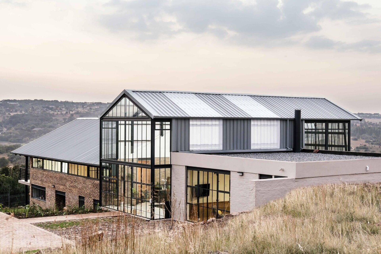 Cement-washed brick, steel, and concrete create a low-maintenance exterior with a strong, modern edge. A double-height conservatory with openable walls and a glass floor becomes the heart of this light-filled home.